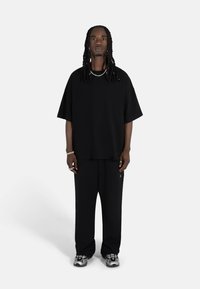 Svart oversized t-skjorte med korte ermer, satt sammen med løstsittende svarte bukser. Brukeren har på seg sølvfargede joggesko og smykker. Enkelt, monokrom design.