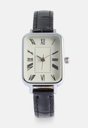 Montre rectangulaire en métal argenté avec un cadran crème, des chiffres romains noirs, un bracelet en cuir noir et une petite couronne sur le côté.