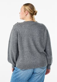 Pull en maille gris avec un col rond, des poignets et un ourlet côtelés. Présente une finition texturée et des accessoires décoratifs sur l'épaule.