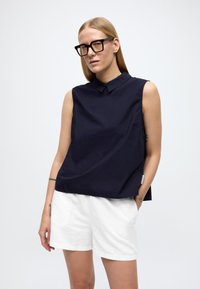 Naine, kellel on sirged blondid juuksed, kandmas suuri musti prille, varrukateta tumelilla tuba ja valgeid lühikesi pükse, seisab ühe käega taskus üheljalgsel lihtsal taustal.