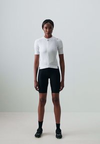 Weißes kurzärmeliges Radtrikot mit Reißverschluss und schwarze Radsportshorts. Kombiniert mit schwarzen Socken und Fahrradschuhen. Raum mit einfacher heller Hintergrund.