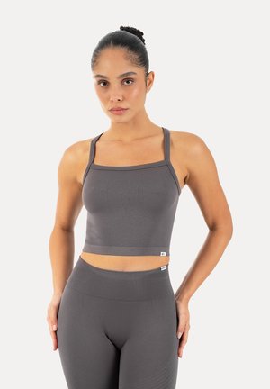 Frau trägt ein passendes graues, eng anliegendes nahtloses Croptop und hoch taillierte Leggings, steht mit einer Hand in der Hüfte vor weißem Hintergrund.