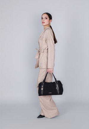 Beige op maat gemaakte pak met wijde broekspijpen en een gebelt jacket, gecombineerd met een zwarte handtas met gouden accenten en ritsen.