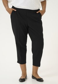 Pantalons noirs ajustés avec une texture lisse, ourlet court et poches latérales, associés à des chaussures plates noires.