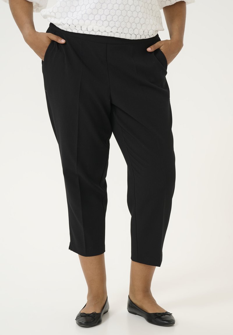 Pantalons noirs ajustés avec une texture lisse, ourlet court et poches latérales, associés à des chaussures plates noires.