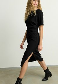 Robe noire ajustée à manches courtes, dotée d'une longueur mi-mollet et d'une fente latérale. Tissu lisse et silhouette près du corps.