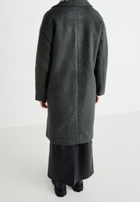 Abrigo gris de mezcla de lana con un corte relajado, mangas largas y un detalle de costura en la parte posterior, combinado con una falda negra de aspecto cuero y zapatos gruesos.