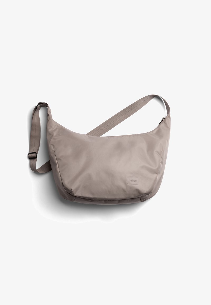 Sac bandoulière marron clair en forme de croissant avec sangle réglable et logo discret de la marque à l'avant, isolé sur fond blanc.