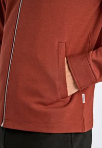 Röd zip-up hoodie gjord av texturerat tyg, med en sidficka och en liten logotypetikett på fållen. Designen har en avslappnad passform.