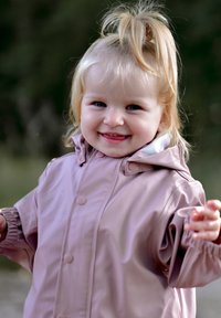 Šviesiai rožinė neperšlampama striukė su gobtuvu, turinti sagų užsegimus ir elastines rankoves, dėvima besišypsančio jauno vaiko lauke.