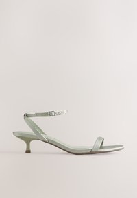 Sandale argentée à petit talon avec fine bride à la cheville et boucle, bout ouvert et design minimaliste sur fond neutre.