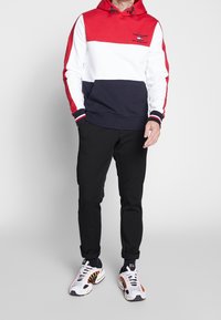 Czerwony, biały i granatowy kolorowy bluz z kapturem z regulowanym kapturem, ściągaczami przy mankietach oraz kieszenią typu kangur, w połączeniu z czarnymi spodniami i białymi sneakersami.