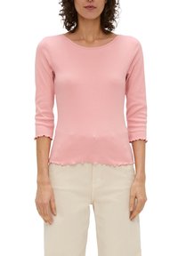Vrouw in een aansluitend roze geribbeld shirt met driekwart mouwen en een gekartelde zoom, gecombineerd met beige hoge taille broek.