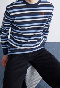 Sininen, laivastonsininen ja valkoinen raidallinen pitkähihainen paita, jossa on pyöreä kaula-aukko. Paidan etuosassa on PUMA-logo oranssina. Mustat farkut viimeistelevät asun.