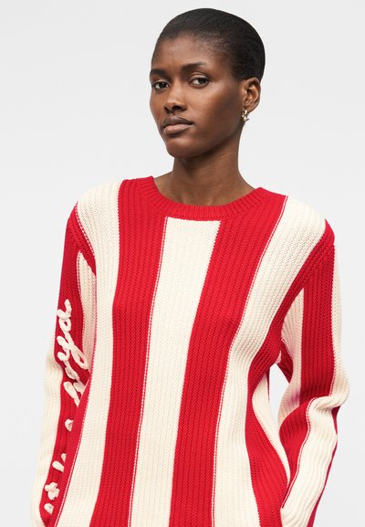 Γυναίκα με κοντά μαλλιά που φοράει κόκκινο και άσπρο κάθετα ριγέ πλεκτό πουλόβερ με κείμενο στο αριστερό μανίκι, σε απλό φόντο.