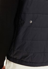 Svart vadderad väst med horisontella sömmar, med tryckknappslåsning och elastisk nederkant. Tillverkad av ett mjukt, lätt material.