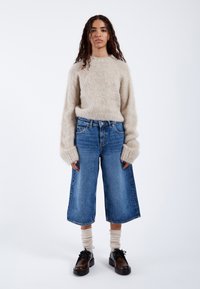 Beżowy puszysty sweter z długimi rękawami, w połączeniu z szerokimi niebieskimi dżinsowymi kulotami. Model ma na sobie ciemne buty i jasne skarpetki.
