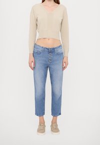 Mujer con un suéter corto beige de cuello en V, jeans azules de tiro alto con botones, y zapatillas beige, de pie contra un fondo blanco.