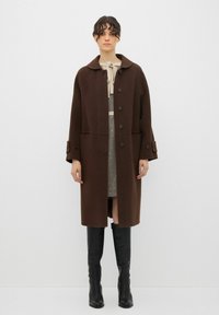 Cappotto di lana marrone con un ampio colletto, sei bottoni e grandi polsini, abbinato a una gonna fantasia e stivali neri al ginocchio.
