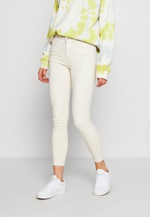 Person iført off-white skinny jeans, en gul-grøn tie-dye sweatshirt og hvide sneakers, stående mod en ensfarvet lys baggrund.