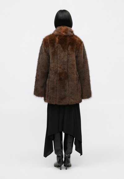 Abrigo de piel sintética marrón con un alto cuello, superficie texturizada y costuras verticales, llevado sobre una falda negra larga y botas negras hasta la rodilla.