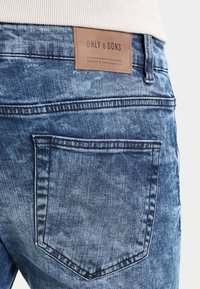 Jeans in denim blu con un finish usurato, caratterizzati da una toppa in pelle marrone etichettata "ONLY & SONS" sulla vita e in una tasca posteriore.