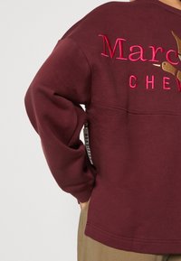 Sweat-shirt bordeaux avec texte brodé rose et or "Marc Chevignon" dans le dos, porté par une personne ayant la main dans la poche.