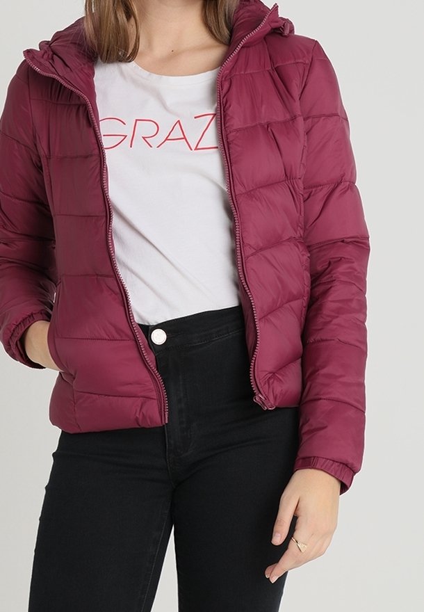 Giacca a vento bordeaux con cappuccio, chiusura con zip e pannelli cuciti. Abbinata a una t-shirt bianca con scritte rosse e pantaloni neri.