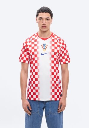 Mlad moški, oblečen v rdeče-belo karirasto hrvaško nogometno majico in modre kavbojke, stoji pred enobarvnim ozadjem.