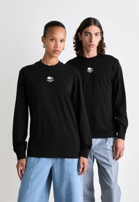 Zwei Modelle tragen schwarze Langarmhemden von Lacoste Paris; eines kombiniert mit hellblauen weit geschnittenen Jeans und das andere mit gestreiften hellen Hosen.
