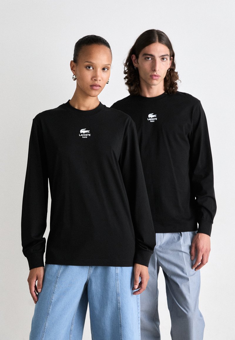 Zwei Modelle tragen schwarze Langarmhemden von Lacoste Paris; eines kombiniert mit hellblauen weit geschnittenen Jeans und das andere mit gestreiften hellen Hosen.