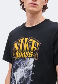 Jeune personne portant un t-shirt noir avec un grand texte jaune ""Nike hoops"" et une illustration d'un panier de basket et d'un filet.
