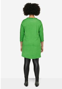 Tunique en lin vert à manches trois-quarts, avec un ourlet arrondi et des bords effilochés, associée à des leggings noirs et des bottes à chevilles cloutées.