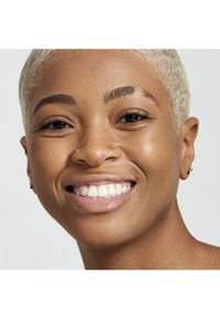 Nahaufnahme einer Person mit hellblondem Haar, klarer Haut und breitem Lächeln, mit sichtbaren Nasenpiercings und dezenten Sommersprossen.