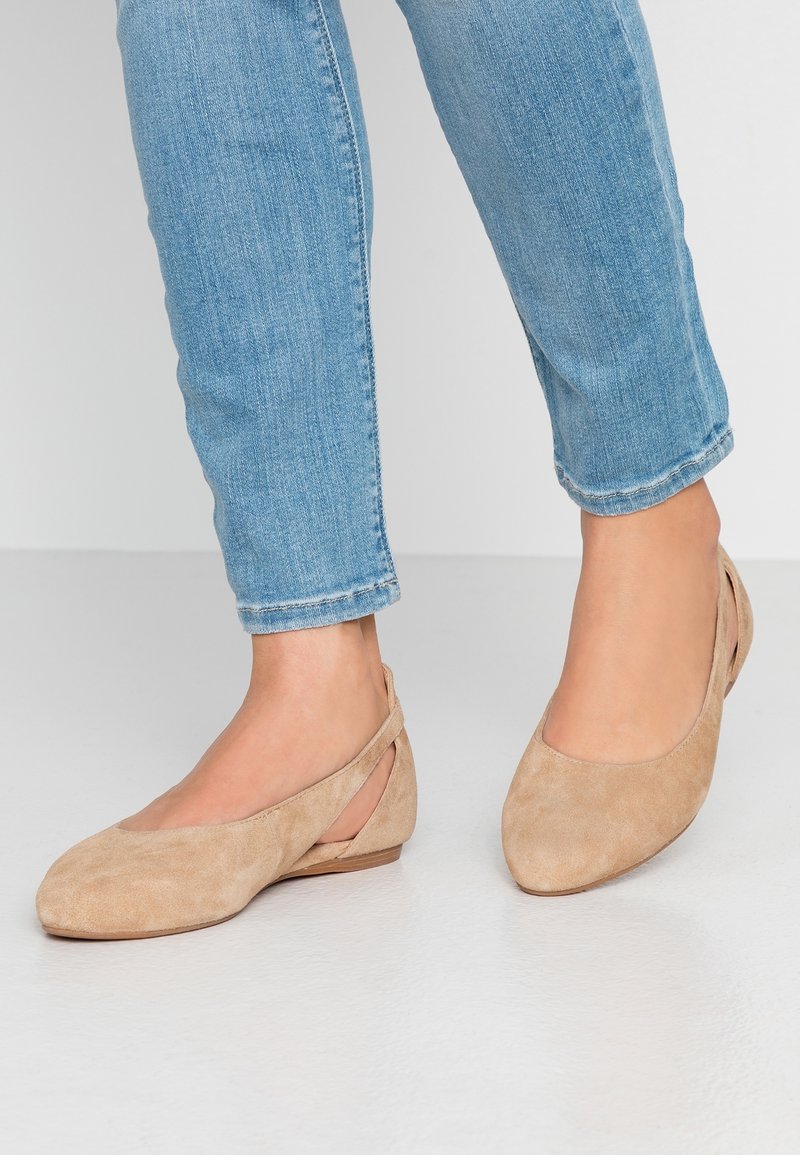 Pieds portant un jean bleu clair et des chaussures plates en daim beige avec des brides ouvertes à l'arrière, debout sur un sol blanc.