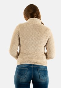Giacca in pile beige morbido con superficie texturizzata, colletto alto e design aderente, abbinata a jeans scuri aderenti blu.