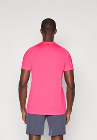 Αθλητικό μπλουζάκι με κοντά μανίκια σε φωτεινό ροζ χρώμα, με στρογγυλή λαιμόκοψη και ανακλαστική λεπτομέρεια στο πάνω μέρος της πλάτης. Φορεμένο με γκρι σορτς.