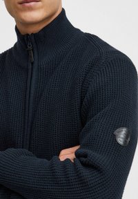 Temno plavača zip majica s teksturiranim pletenim dizajnom, visokim ovratnikom i okruglim crnim logom na lijevom rukavu.
