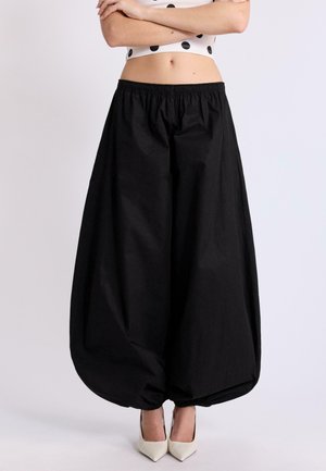 Osoba nosiaca čierne balónové nohavice so širokými nohavicami a elastickými lemami, doplnené bielymi špicatými vysokými podpätkami a bielym krátkym topom s čiernymi bodkami.