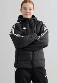 Svart pufferjakke med hette, med hvite striper langs skuldrene, glidelås og to sidelommer. Strukturert, quiltet materiale.