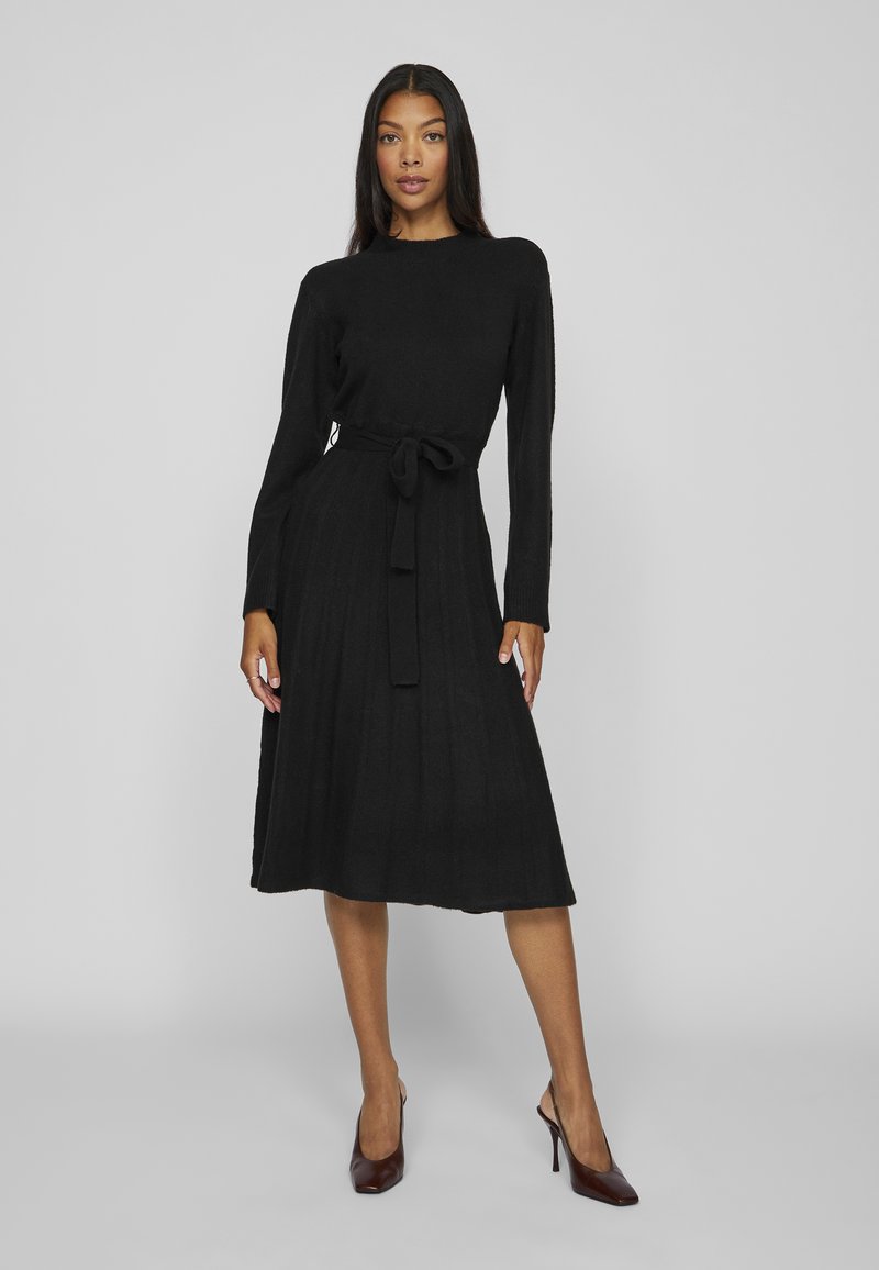 Black knitted dress with long sleeves, ribbed texture, knee-length, and a fabric belt tied at the waist. Brown high-heeled shoes complete the look.
