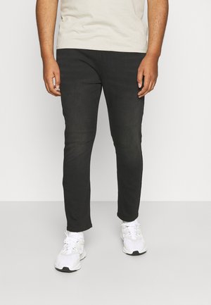 Mann trägt schwarze Slim-Fit-Hose, weiße Sneakers und ein beiges T-Shirt, steht vor einem schlichten Hintergrund.