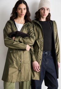Two individuals wear olive green waterproof jackets with zippers and pockets. One holds a dark brown fleece, while the other wears a black shirt and blue jeans.