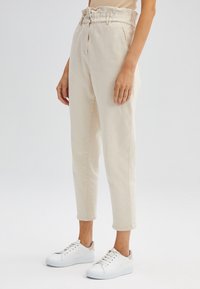 Kvinna i högmidjade beige byxor med uppvikt fåll och vita sneakers, som står mot en slät ljus bakgrund.