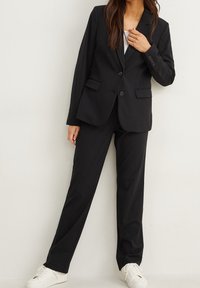 Femme portant un costume noir sur mesure avec un haut blanc et des baskets blanches, se tenant devant un mur clair uni.