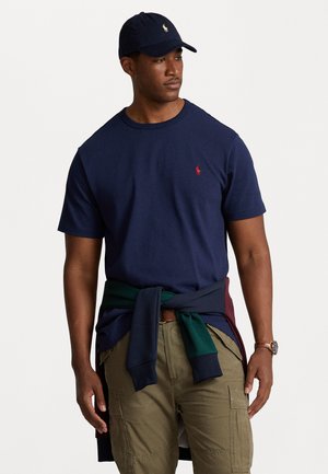 T-shirt en coton bleu marine avec un petit logo rouge sur la poitrine, associé à un pantalon cargo olive et un pull vert et bordeaux noué à la taille.
