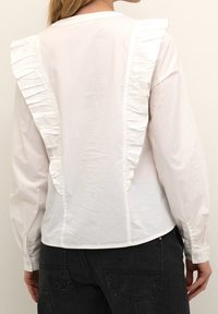 White long-sleeve shirt with ruffled accents down the back, featuring a smooth texture and buttoned cuffs. Paired with dark jeans.