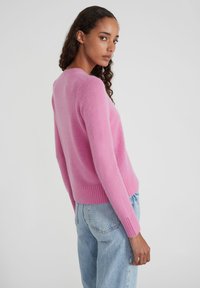 Frau mit lockigem Haar, die einen rosa Pullover und hellblaue Jeans trägt, seitlich steht und über ihre Schulter blickt, vor einem einfarbigen Hintergrund.
