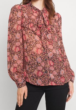Vrouw die een doorschijnende bloemenblouse draagt in roze en paarse tinten met een gerimpelde voorkant en lange pofmouwen, gecombineerd met een zwarte broek.