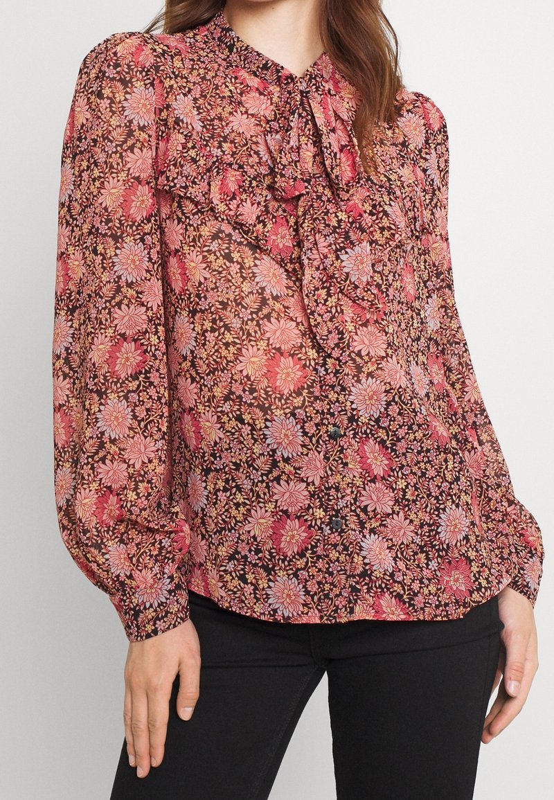 Femme portant un chemisier transparent à motifs floraux dans des tons roses et violets avec un devant à volants et de longues manches bouffantes, associé à un pantalon noir.